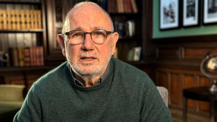 Jimmy McGovern is sitting in a library. A number of books can be seen on shelves in the background. He is wearing a green jumper and glasses and is staring at the camera.