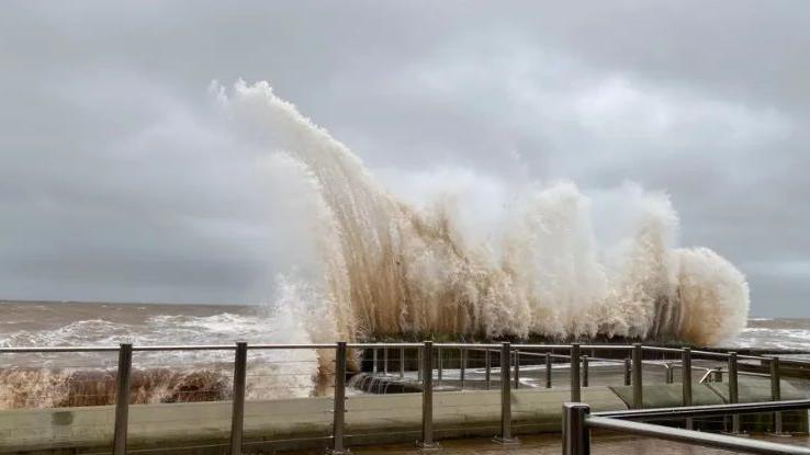 A high wave of muddy brown seawater rises above a quay after hitting it on a grey day. There are safety railings on the edge of the quay.