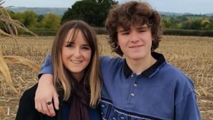 Family photo of Crystal (on the left) and Harvey. Harvey is on the right and has his arm around his mum's shoulders. He's wearing a blue long-sleeved rugby top with a black collar and black stripes across the chest. Crystal is wearing a black coat and a brown scarf. They're both smiling and looking towards the camera, and are stood in a stubbly crop field, with trees and hills visible on the horizon. 