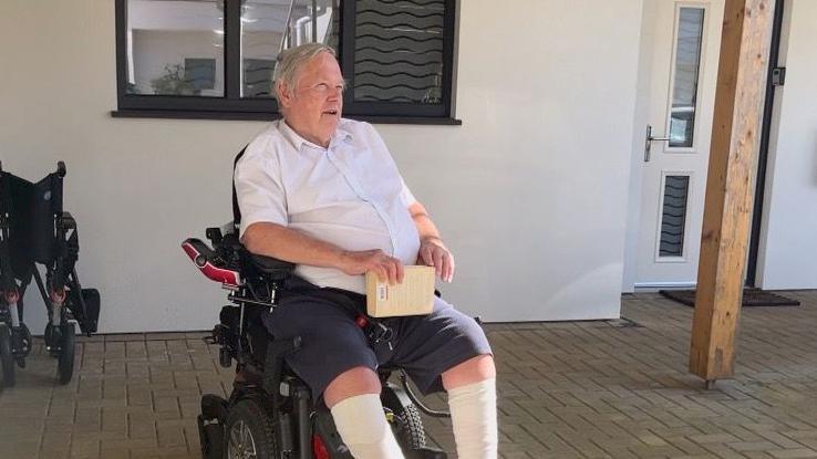 A man in a wheelchair in blue shorts and white shirt holding a book outside a house.