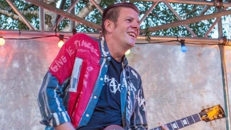 Dave Young pictured on stage playing guitar. He has short brown hair and is wearing a blue t-shirt with a red and blue jacket. He is not looking at the camera, but out to the crowd. he is smiling.