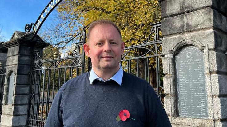 Cllr Davis pictured smiling slightly and looking into the camera. He's wearing a light blue collared shirt, a navy blue woollen jumper, and has a paper poppy pinned to his chest. He's stood in front of huge, wrought iron and stone gates - you can see there's writing over the top of the gates that reads 'Oakengates.' There are trees in the background with yellow leaves, and the sky is blue. 