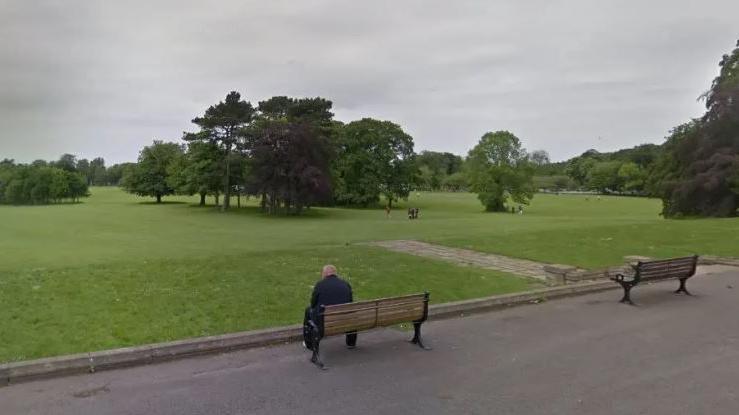 A park, with trees surrounding a grassed area, and a clump of trees in the middle distance. At the front are two seats in a paved or tarmacked area, with a man in a dark coat sitting in one of them.