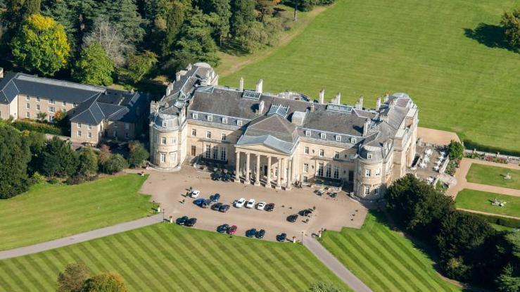 A view of Luton Hoo from above. A luxury stately home used as a hotel. Cars are parked outside the entrance which has six columns in front of it. The lawn in front has mower stripes. There is a large grassy area and woodland behind the house.