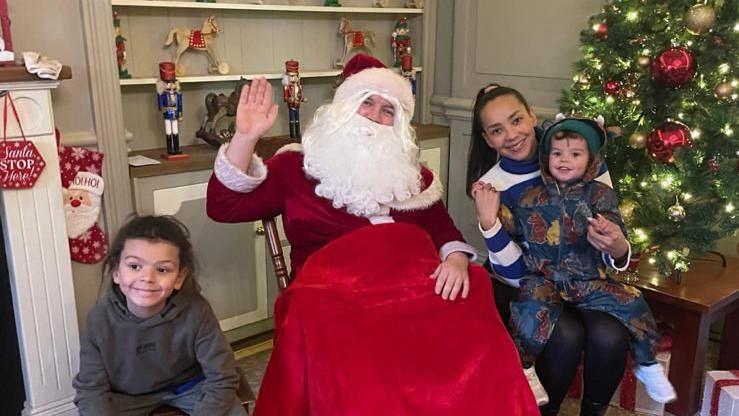 Two young boys and their mum sit next to a man dressed as Santa Claus. Smiles all round and a nice Christmas tree.