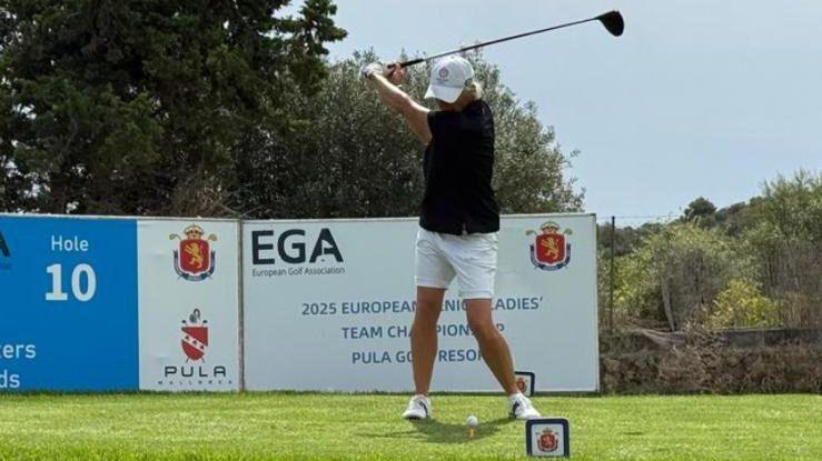 Sally Shayler is captured mid-swing on a sunny course, wearing a black shirt, white shorts, and a white cap. Behind her stands a large event sign that reads “2025 European Men’s and Ladies’ Team Championship” at Pula Golf Resort. The sign marks hole number 10 and displays logos of sponsors. Trees and shrubs frame the background.