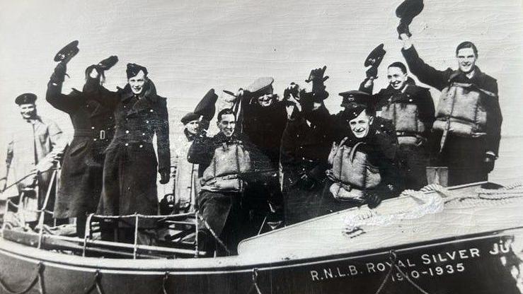 A black and white photo of the RNLI crews from WW2. They are in a lifeboat raising their hats in the air to wave.