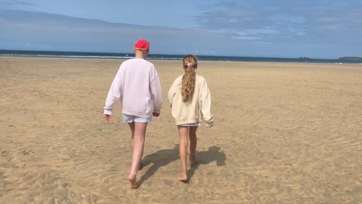 The two girls walking with their backs to the camera on the beach. There is sea and sky in the background.