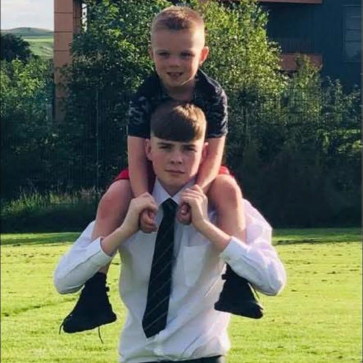A younger child sits on the shoulders of an older teenager standing on a grassy field. The older teenager is dressed in a white shirt, dark tie and black trousers, while the younger child wears a dark top and shorts.