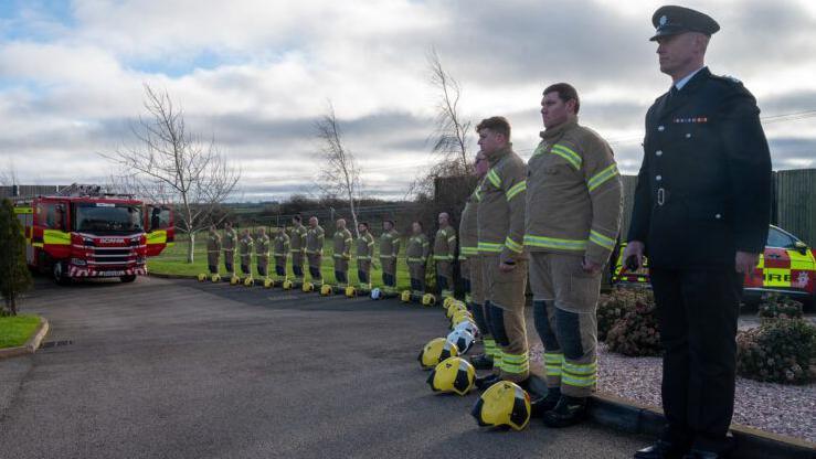 Colleagues line up to honour Gary at the funeral, outside the crematorium. Crews in uniform line the road on the right, with their yellow helmets on the floor in front of their boots. A red fire engine is in the distance. A man wearing a more formal black uniform stands in the foreground.