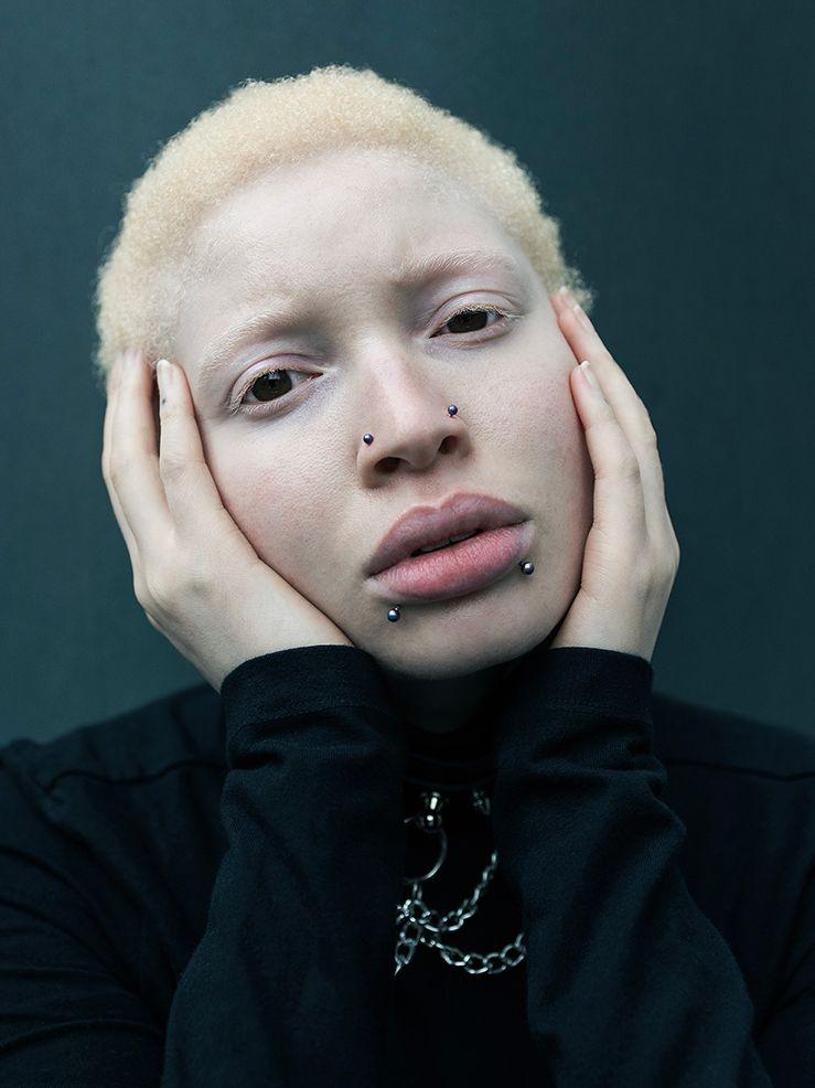 A person with short, light blonde hair and pale skin, wearing a black jumper and silver chains. They have face peircings in their nose and lower lip, and have their head propped up on their hands. They look at the camera with a vacant expression.