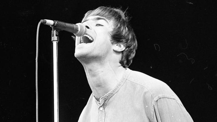 Liam Gallagher in a black and white photograph, he is singing into a microphone.