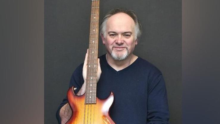 Nick Smith is looking at the camera and smiling. He has dark hair which is grey at the sides and he has a grey beard. He is holding an electric bass guitar and is wearing a blue pullover.