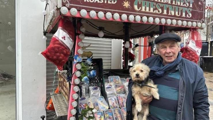 A man wearing a black flat cap holds a dog in his arms while standing in front of his stall.
