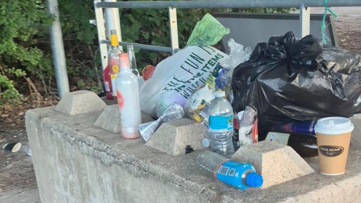 Rubbish bags, used plastic bottles and glass bottles and coffee cups, are left on a concrete barrier on a pathway.