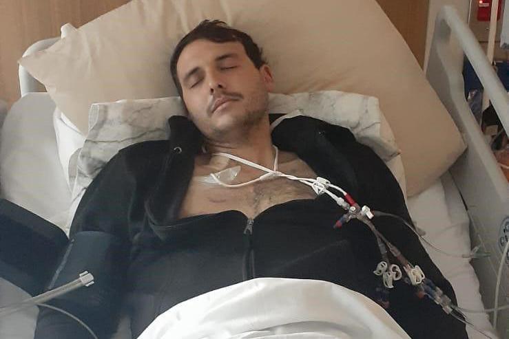 A man lies in a hospital bed with his eyes closed and tubes attached to his chest. He is wearing a black hoodie, open at the chest. A white blanket covers the lower part of his body.