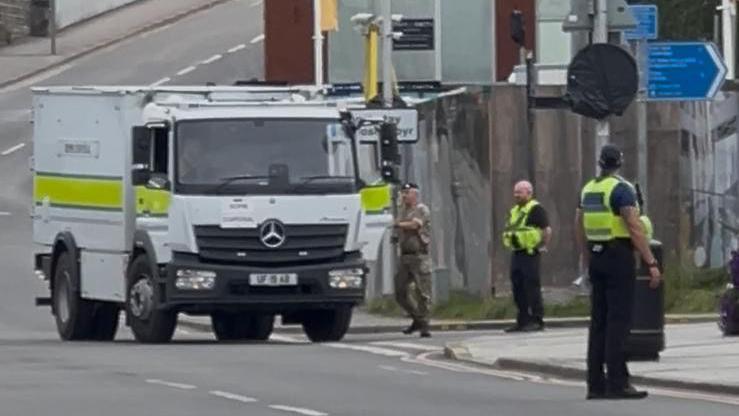 A large white truck on a street in Bridgend. It has a yellow stripe and man in military fatigues standing at the door. Two police officers are also on the street