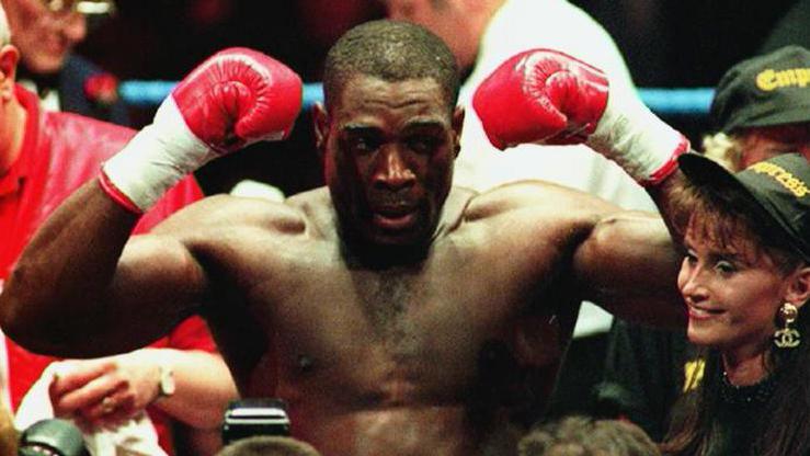 Frank Bruno stands in a ring in the 1990s with his arms raised in triumph, wearing red boxing gloves. 