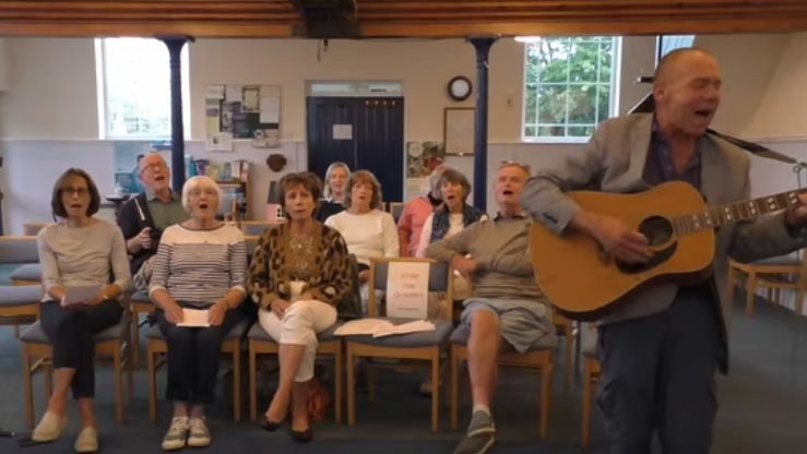 A group of men and women sitting on chair while singing along with a man who is standing up and playing a guitar.