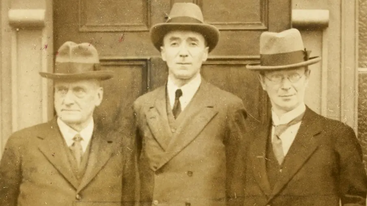 A black and white archive photo of Joseph Fisher, Richard Feetham and Eoin MacNeill during a meeting of the Boundary Commission in Armagh in 1924.  They are all wearing suits. ties and hats and are standing in front of a door.