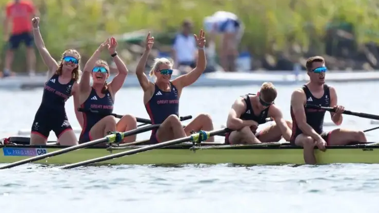 Four rowers and their cox - Erin  celebrate in their boat after winning their final at the Paris 2024 Paralympics. The crew of three women have their arms aloft, while the two men at the rear of the boat are facing downwards as if recovering from their efforts.