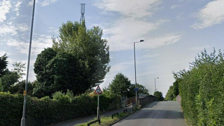 A road bridge with a footpath alongside it. There is a hedge on the right hand side of the image and to the left hand side is another hedge with trees behind it.