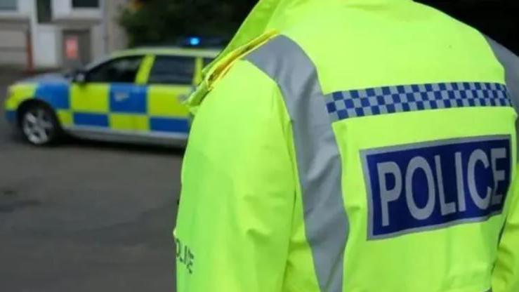 Police officer, photographed from behind, wears a hi-vis jacket. A police patrol vehicle is behind the officer.