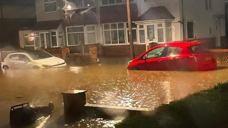 Two parked cars wheel-deep in a flooded road where water has risen and reached the walls of semi-detached houses and knocked over a wheelie bin