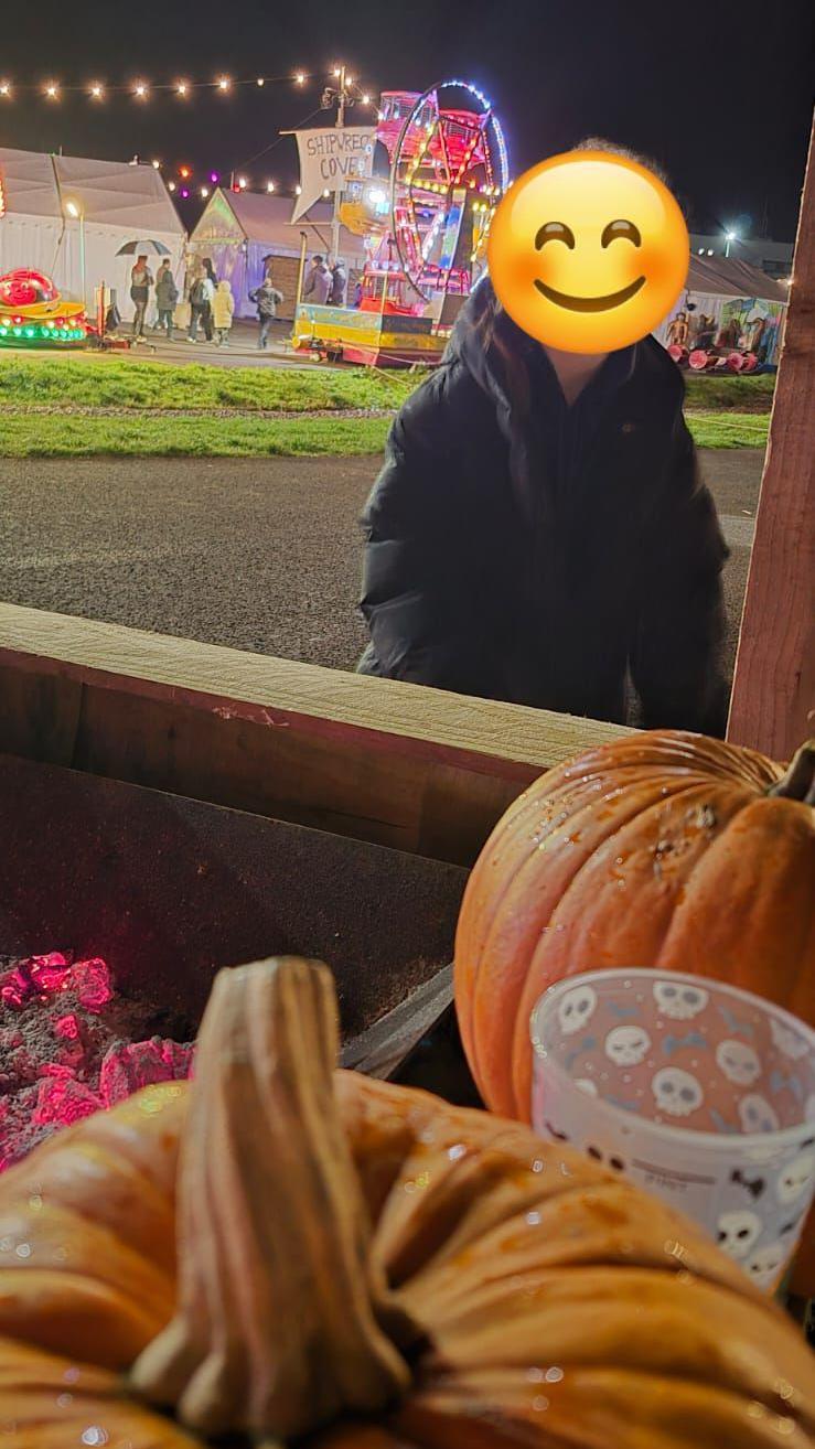 A child with a face hidden by a smiling emoji stands in front of some pumpkins. Behind is what looks like a wooden pirate ship, two small fair rides and a number of white marquees. There is an area with tarmac and strips of grass behind the child.