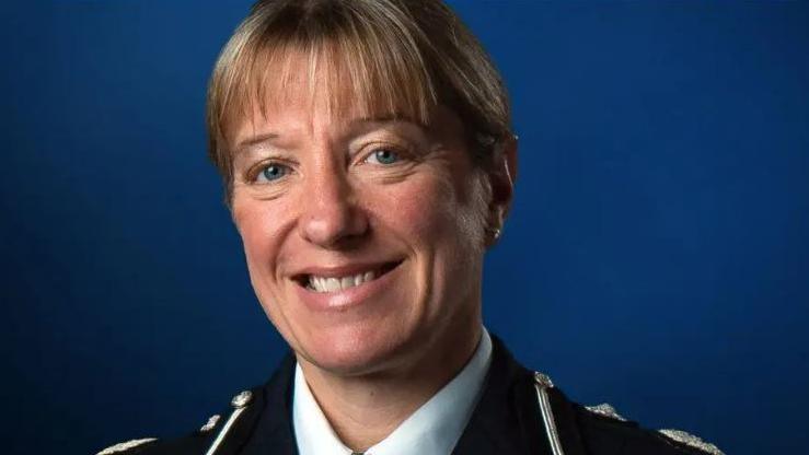 A blonde woman in a police uniform smiles at the camera in front of a dark blue backdrop