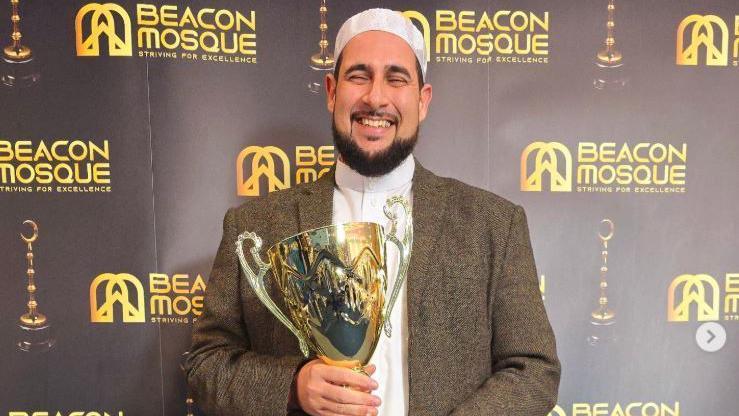 Imam Adam Kelwick who has a dark brown beard has his eyes shut as he has a huge smile holding a trophy at the Beacon Mosque Awards 