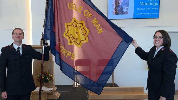 Dylan and Rachael Nieuwoudt in uniform holding up a red and blue flag with the words Isle of Man Corps written on it with a yellow octagon in the middle featuring the words Blood & Fire in red writing. Wooden church fixtures including chairs, a lectern and an arrangement of flowers can be seen in the background.