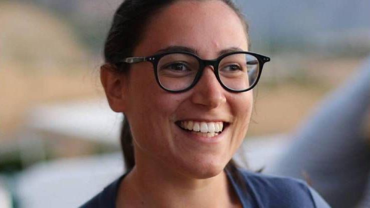 Sofia Brizio, a young woman with brown hair, brown eyes, wearing dark framed glasses and wearing a mid blue top smiles looking slightly off camera, the background is blurred.