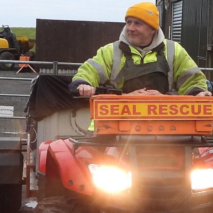 Phil is riding a red quad bike. The machine's lights are on and it has an orange sign on the front with the words "seal rescue" in orange letters on a yellow background. The quad bike is pulling a small trailer with a sheet of black tarpaulin over the top of it. Phil is wearing green waterproof overalls over a yellow safety jacket. He is wearing a bright orange, woolly hat.