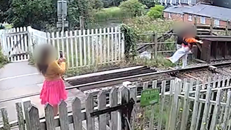 CCTV footage of a railway line foot crossing. A person wearing a yellow top and pink skirt, with long brown hair is holding their phone up to take a photo of someone standing beside the railway line