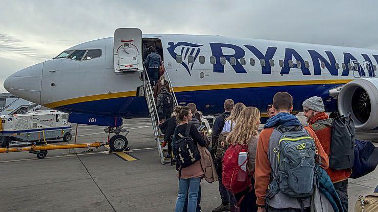 Passengers - mainly aged between 18-35 - boarding a Ryanair aircraft near the cockpit, via a mobile stairway on an airport tarmac under a cloudy sky. The aircraft is white with a blue and yellow stripe and the word ‘RYANAIR’ prominently displayed on the fuselage. People in casual clothing with backpacks and luggage are lined up, walking toward the plane’s front entrance.