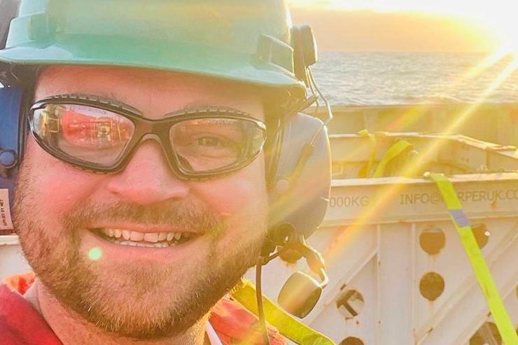 Offshore worker Phil Perry in hard hat, smiling at camera on an offshore platform, with sea and sunshine behind him.