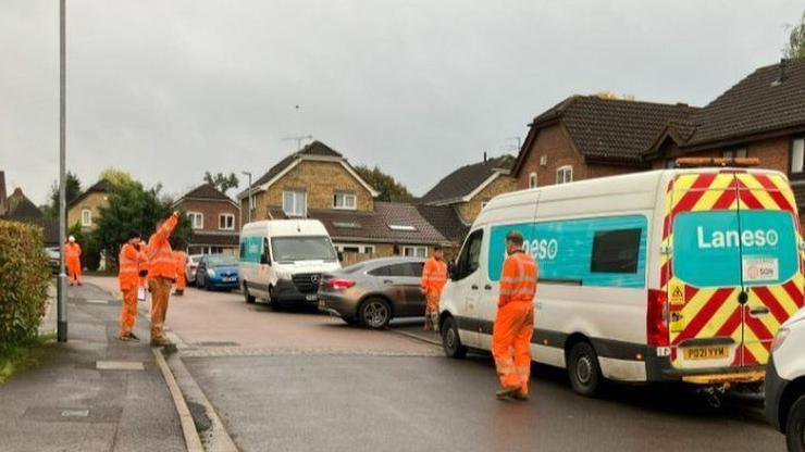 Vans line right hand side of residential street with workers in orange on pavement and in road with homes seen in background