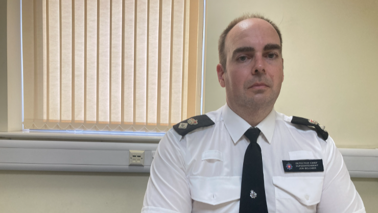 Jon Belcher is sat at his desk in uniform with a window with blinds behind him. He is looking at the camera, with a fairly serious expression. He has short brown hair.