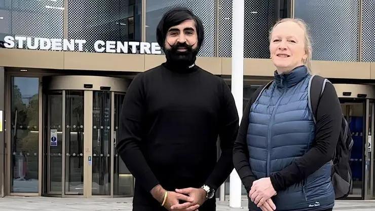 A man with long dark hair and a curled up moustache stands next to a woman with long blond hair. They are outside a university building with a sign that says 'student centre'. 