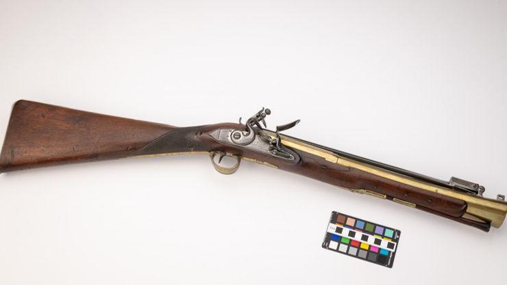 A blunderbuss gun is laid out on display in this image. It has a long wide wooden handle with a long flared brass barrel, a brass trigger guard and an iron trigger and lock.