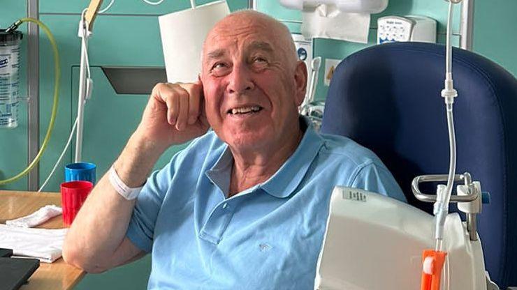 A bald man wearing a blue t-shirt sitting in a hospital chair. There are various bits of medical equipment and machines surrounding him.