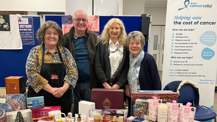 A group of smiling people stand in front of a table full of toiletries and other gifts, with a banner besides them with information about Jersey Cancer Relief