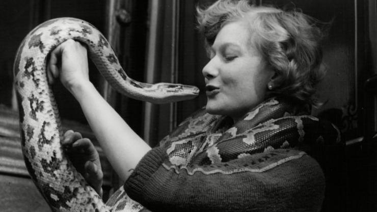 A black and white picture of a woman holding a snake, which is coiled around her and is close to her lips as if to suggest it is going in for a kiss.