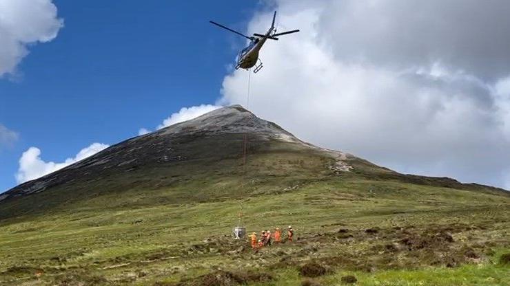 In the foreground a helicopter hovers over the slopes of a small mountain - rocks are suspended from it by a cable