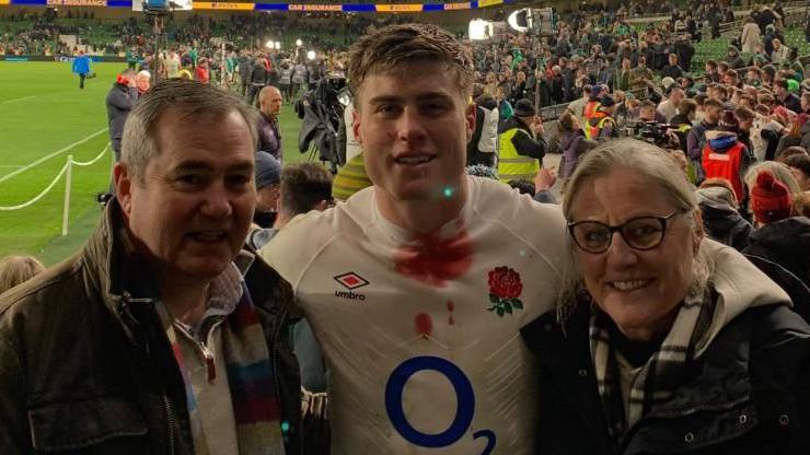 From left-to-right, Cliff Freeman with short grey hair wearing a brown jacket, Tommy Freeman with short dark hair wearing a white England shirt with an O2 logo on the front, and Sara Freeman with short light-coloured hair and glasses wearing a dark jacket with white hood. Tommy has his arms around them and they are standing in a sports stadium with a crowd of people behind them. The rugby pitch is visible to the left.