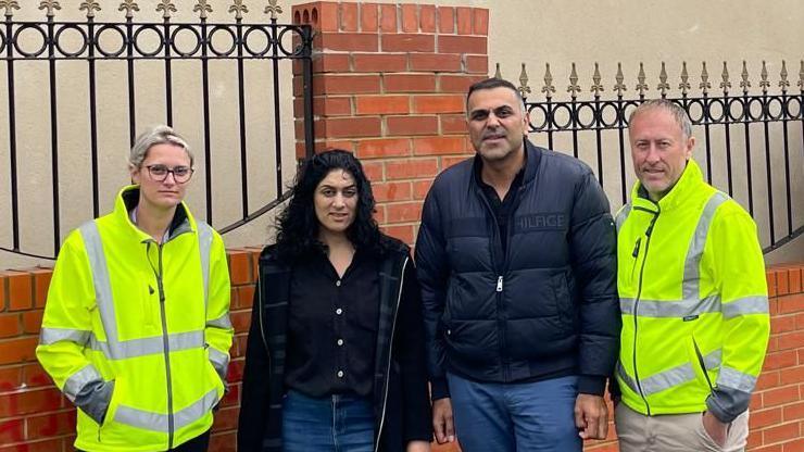 Saima and Husman Rafiq are wearing black coats and standing in front of a brick wall. They look serious and are flanked by two members of council staff, who are wearing hi-vis coats.
