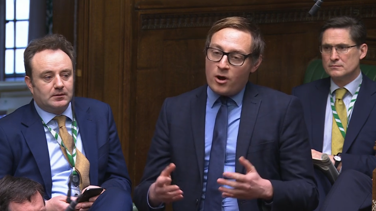 Olly Glover gives a speech in parliament. He is wearing a blue shirt, blue tie and blue suit and is surrounded by other MPs.