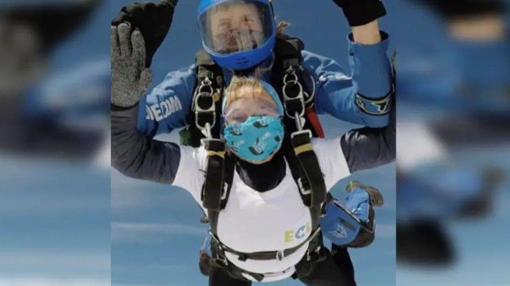 A man and woman tied together jumping out of a plane with their hands in the air and wearing goggles and facemasks with blue skies in the background.
