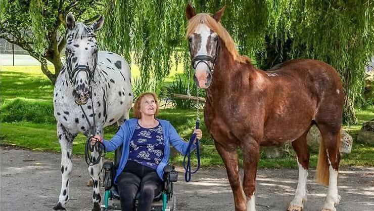 Anne Dunham is sitting in a wheelchair next to two horses. She is looking up at one of the horses and is smiling. Behind them are trees and the sun is shining.